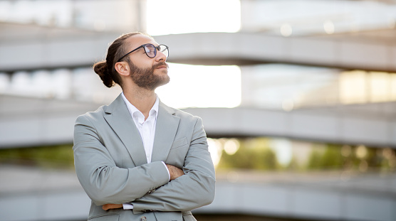 Confident young businessman in suit standing outdoors and looking upwards, handsome male entrepreneur with crossed arms embodying vision and ambition, posing against modern architectural backdrop
