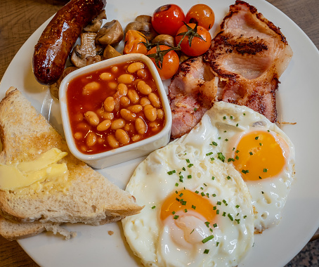 Close up of a full plate of English Breakfast with eggs, bacon, sausage, baked beans, mushrooms, cherry tomatoes and buttered bread.