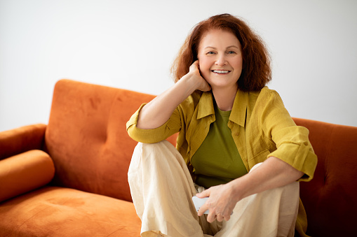 Beautiful Senior Woman With Smartphone In Hand Relaxing On Comfortable Couch At Home, Happy Redhead Elderly Female Resting In Living Room Interior, Enjoying Retirement Time, Smiling At Camera