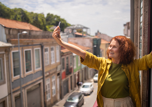 Joyful senior woman stretching out her arm and taking selfie on balcony with urban street view, smiling elderly female using smartphone, capturing photos at home, posing at cellphone camera