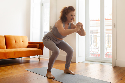 Smiling senior woman making squat exercise at home, active sporty elderly lady training on her yoga mat, smiling during fitness workout in her airy, well-lit living room, copy space