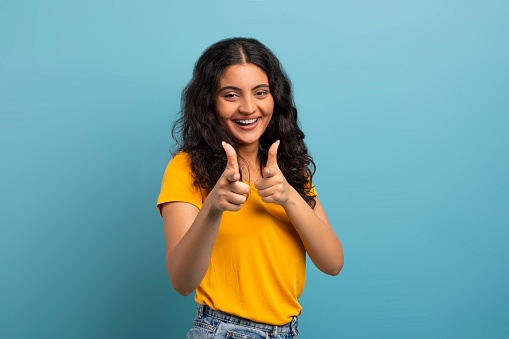 Positive young indian lady student pointing with both hands at camera and smiling, posing isolated on colorful blue studio background, copy space. Gestures concept