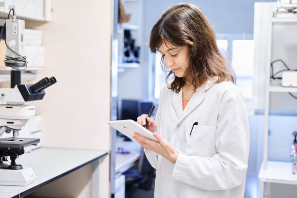 istock Young woman biologist in white coat standing near a microscope and taking notes with a stylus on a tablet during research work in the laboratory. 2059055606