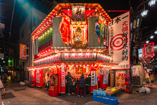 osaka, japan - dec 04 2023: Illuminated shooting game attraction store in the Tsutenkaku Hondori Shopping Street of Shinsekai district adorned with a golden statue of Biliken and ykai demon at night.