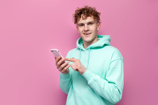 young curly guy in mint hoodie uses smartphone on pink isolated background, man in blue sweatshirt clicks on the screen and chooses online in the phone