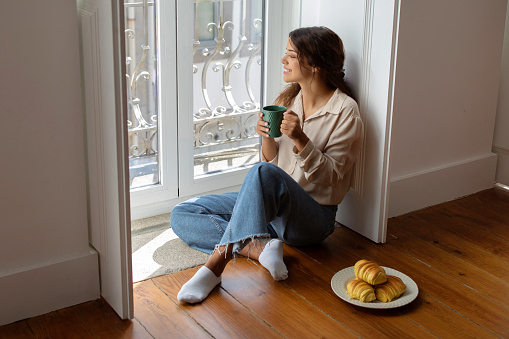Smiling young woman enjoying her morning coffee, sitting on floor by bright window with croissants, happy dreamy millennial lady exuding peaceful weekend vibe in cozy home setting, free space