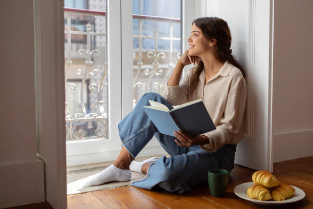 istock Young woman reading book while sitting by sunny window with a view 2059090925
