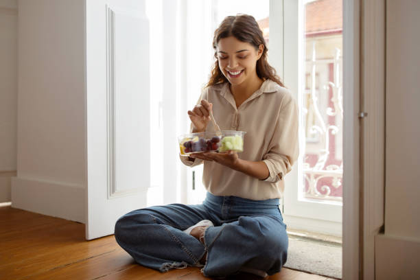 istock Young woman enjoying healthy fruit salad while sitting on floor by window 2059090941