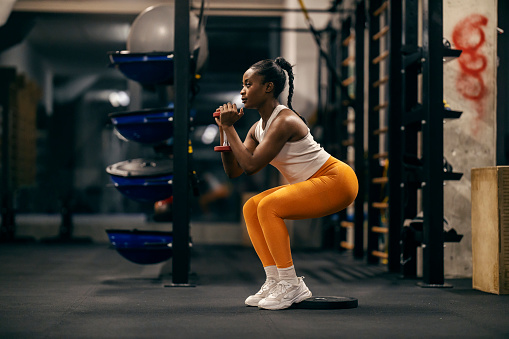 Profile view of a fit black sportswoman practicing squats and holding dumbbells at gym.