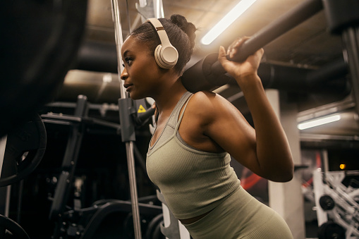 A strong african american sportswoman with headphones doing workout on smith machine at gym and squatting.