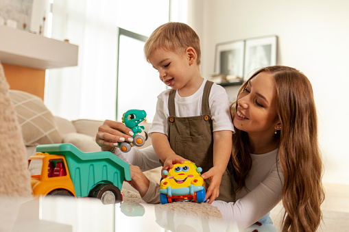 young mother plays with her little son with toys at home and smiles, 2-year-old boy plays with toy car with his parent, woman has fun with her child
