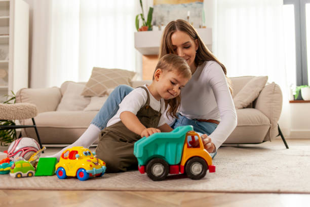 young mother plays with her little son with toys at home and smiles, 2-year-old boy plays with toy car with his parent young mother plays with her little son with toys at home and smiles, 2-year-old boy plays with toy car with his parent, woman has fun with her child child playing with toys stock pictures, royalty-free photos & images