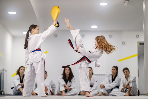 Taekwondo girls practicing kicks with kick pads on training at martial art school. Teammates sitting and watching them.