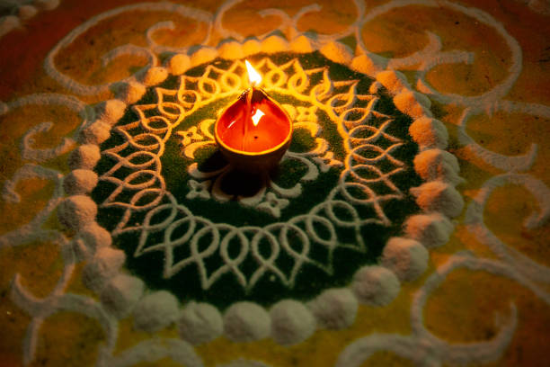 A close-up shot of a woman's hand adorned with bangles delicately decorating a surface with vibrant flower petals and small oil lamps (diyas). The colorful petals and warm glow of diyas create an intricate pattern adds a touch of elegance and festivity. This image captures the essence of tradition and celebration, showcasing the intricate details involved in preparing for a special occasion of Diwali. The woman's gentle touch reflects care and artistry in creating a beautiful atmosphere