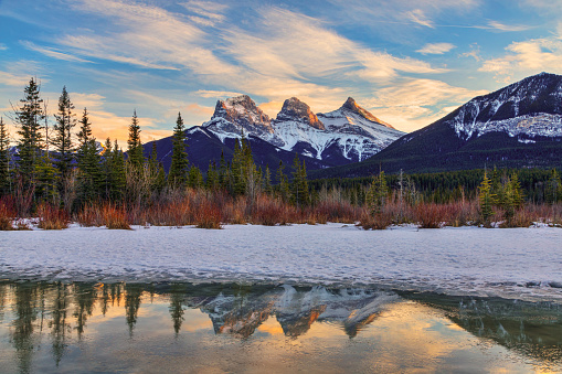 Golden winter sunset at The Three Sisters, a trio of peaks near Canmore, Alberta, Canada, in the Canadian Rockies, with alpenglow on the mountains reflecting off melted snow and ice in the foreground.