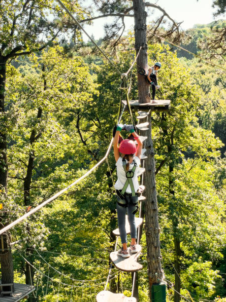 summer fun and sports for adventurous people.happy school girl enjoying activity in a climbing adventure park on a summer day.a teenager girl walks a rope bridge between trees in an amusement park in safety gear and a helmet.rope park in wood forest. - dauntless zdjęcia i obrazy z banku zdjęć