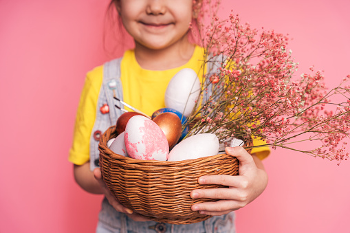 Little girl wearing bunny ears on her head holding basket of Easter eggs in her hands against pink background.