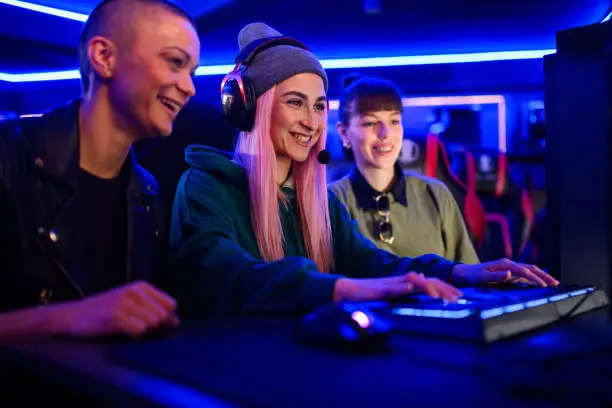 Portrait of a eSports woman gamer with a beanie cap and a pink hair playing online strategy video game on a computer in a internet cafe or cybersport gaming lounge with her female friends.