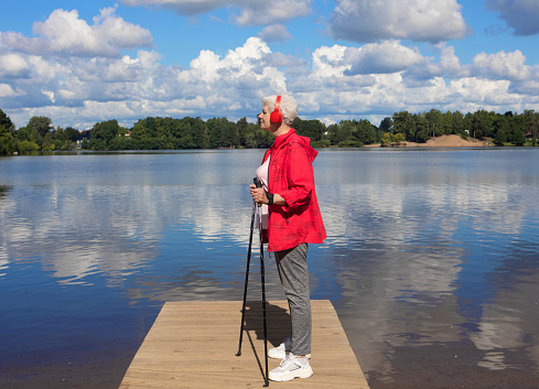 An elderly woman with short hair, dressed in a red jacket, stands with Nordic walking poles on a bridge by the lake nd listening music with headphones.