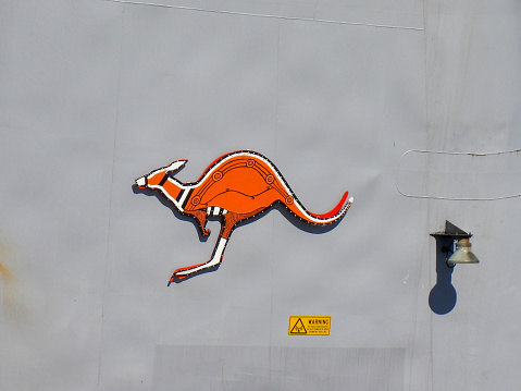 Insignia on the funnel of HMAS Arunta, an Anzac Class frigate of the Royal Australian Navy docked at Garden Island, Sydney Harbour.   The red kangaroo is the national insignia affixed to all major fleet ships and this has been painted in an Indigenous art style.  A warning sign and lighting equipment are also attached to the funnel.  This image was taken from Woolloomooloo Bay on a hot and sunny afternoon on 3 March 2024.