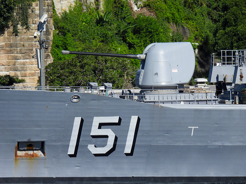 The forward turret and 5 inch Mk45 Mod 2 automatic rapid fire gun of HMAS Arunta, an Anzac Class frigate of the Royal Australian Navy.  She is docked at Garden Island, Sydney Harbour. In the background is a pole with six visible security cameras. This image was taken on a hot and sunny afternoon from Woolloomooloo Bay on 3 March 2024.