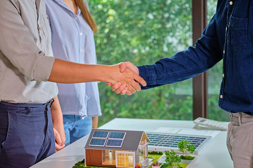 Close up hand couples shaking hands with Solar energy sales engineering  over a  home model  featuring solar panels successful  deal agreement  symbolizing a green energy  sustainable