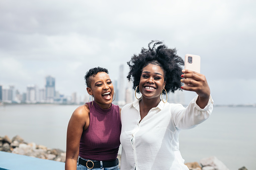 Two stylish friends taking a selfie with their smartphone leaning on a bench next to the sea. In the background you can see the entire skyline of Panama City with all the skyscrapers