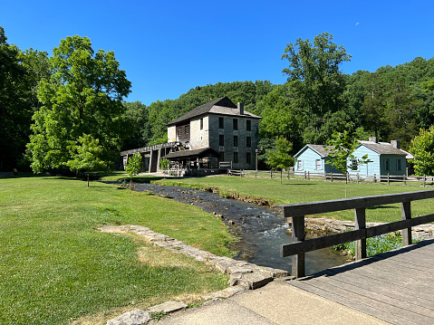 Mitchell, Indiana, USA - June 20, 2022:  Mercantile, Apothecary, Saw Mill and Grist Mill beside a creek with a foot bridge in the recreated and restored 1800 Pioneer Village at Spring Mill State Park, near Mitchell, Indiana with beautiful blue sky copy space and vivid green trees and grass.