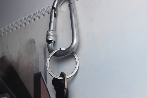 A silver key chain carabiner in the shape of a rope-tying tool used for climbing mountains is attached to the end of a nail on the wall of the house.