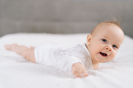 Closeup of funny newborn baby 4 month laying on stomach developing neck control .Tummy time for strengthen baby neck and shoulder muscles. Cute infant lying crawling on white bed happiness and fun