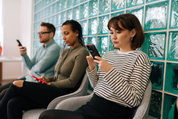 poważni, wielokulturowi młodzi biznesmeni siedzą w rzędzie ze smartfonami, czekają na rozmowę kwalifikacyjną, siedzą w kolejce na krzesłach w nowoczesnym biurze - unemployment waiting in line waiting room interview zdjęcia i obrazy z banku zdjęć