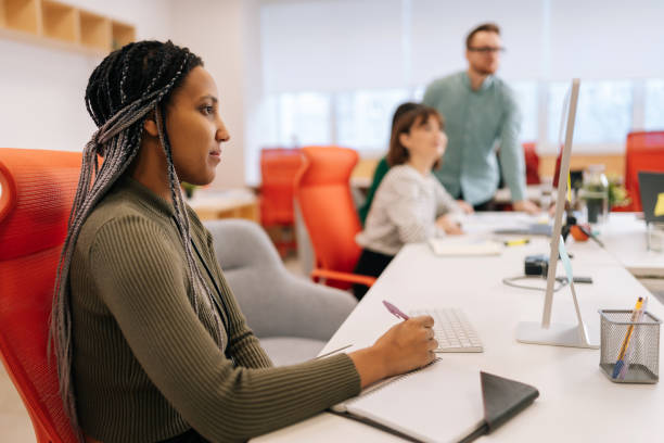 istock Side view of young African woman writing down notes and using computer sitting at work in big modern corporate office. Colleagues businesspeople working together 2059907063