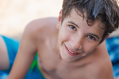 Close-up on cute preadolescent boy in trunks lying on the beach with wet hair and smiling at camera, family vacation in late summer