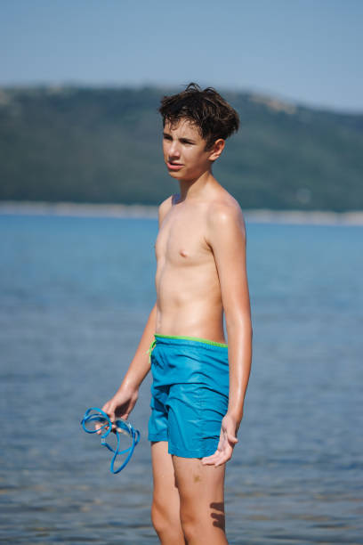 Preadolescent boy in blue swimming trunks standing by the water and holding googles before a swim, family vacation in late summer