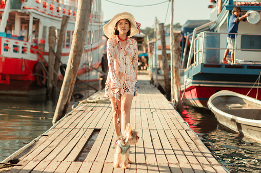 Happy Asian woman in casual clothes Walk with Yorkshire terrier on a wooden bridge in a local fishing village. Enjoy the summer vacation with your family. Surrounded by natural beauty and happiness.