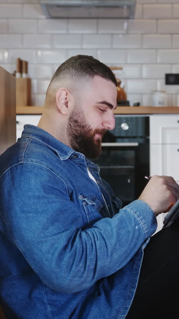 Vertical video. Confident young adult business man sitting on the floor leaning against the kitchen counter working and writing on touch screen of tablet device with tablet pen