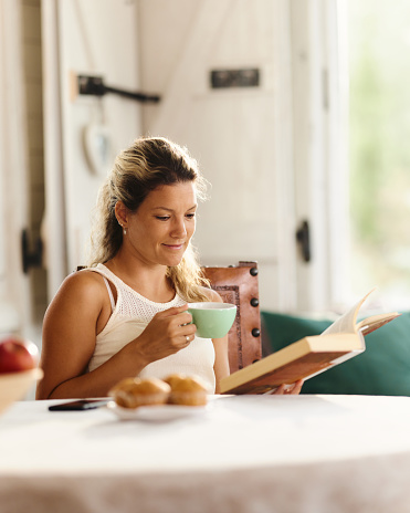 Smiling woman drinking coffee and enjoying in book reading at the table in dining room. Copy space.