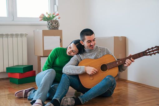 A young couple is enjoying their new apartment, sitting on the floor surrounded by boxes. A man plays the guitar and a woman rests her head on his shoulder.
