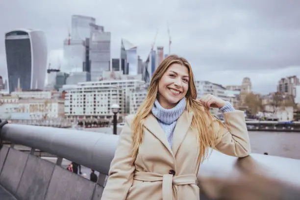 Close up portrait of a young cheerful blond girl in her 30s posing and smiling in toothy smile, while looking directly in camera, the background is city of London's skyline, England, UK. Selective focus with priority on the model and plenty of copy space at the background and sky, which is defocused down town modern corporate business buildings and financial district near Liverpool Street at the heart of the capital. Photo created during cold season outdoors and the model is with warm casual clothes in light cream and blue colours on a cloudy day  - creative stock photo