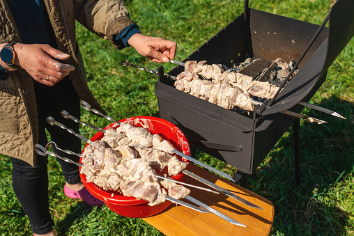 Woman strands raw pieces of marinatedo pork meat on long skewers frying shish on the grill.Nice summer day.