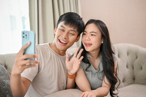Lovely, happy young Asian couple taking selfies or recording a video with a smartphone together while relaxing in the living room. people and wireless technology concepts