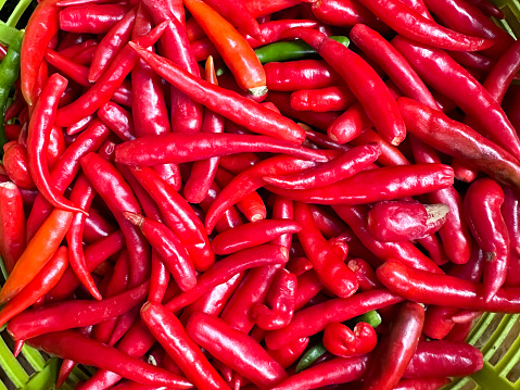 Stock photo showing close-up, elevated view of a heap of glossy red chilli peppers, with green stalks, in a green plastic basket being sold at an outdoor fruit and vegetable market.