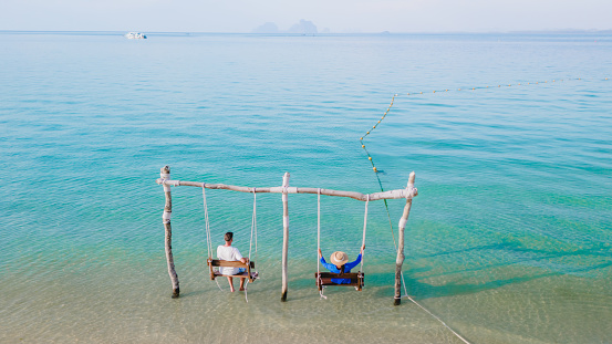 a couple of men and a woman on a swing at the beach of Koh Muk a tropical island with palm trees soft white sand, and a turqouse ocean, Koh Mook Trang Thailand, a couple on a swing in the ocean sunset