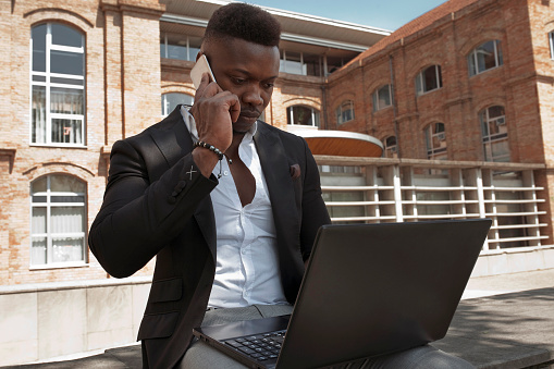 African american businessman works with laptop and smart phone outdoors at city. Manager, business concept.