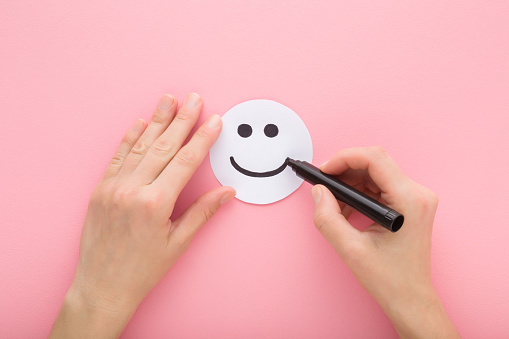 Young adult woman hand holding black marker and drawing happy smiling face on white paper on light pink table background. Pastel color. Closeup. People mood concept. Point of view shot. Top down view.