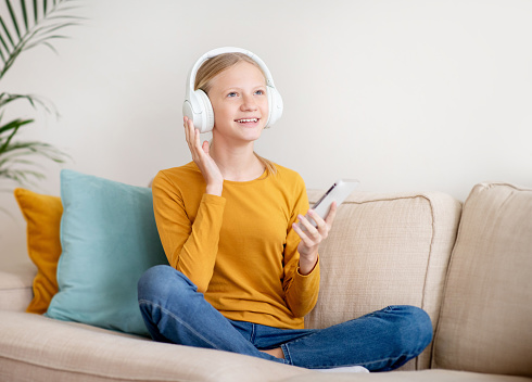 Portrait of happy teen girl wearing wireless headphones listening music on smartphone, cheerful teenage female child enjoying favorite songs while sitting on comfortable couch at home, copy space
