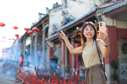 A joyful solo Asian woman traveler captures moments on her smartphone while exploring Georgetown, Penang. Traveling light, she embraces the vibrant streets and colorful buildings, finding joy in simple moments and creating cherished memories along the way.