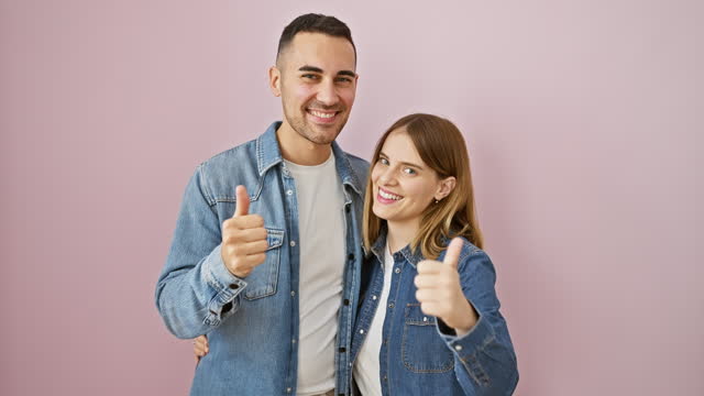 Beautiful couple wearing denim shirts, beaming with success. together they stand, giving cheerful thumbs-up gestures, expressing approval over pink isolated background.