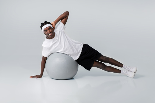 Smiling black man exercising in side plank position on gym ball, motivated young african american guy wearing white t-shirt and headband making core workout against grey studio background, copy space