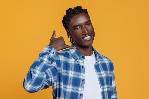 Call Me. Cheerful Young Black Guy With Hand Near Ear Imitating Phone And Looking At Camera, Handsome Millennial African American Man Inviting For Communication While Standing On Yellow Background